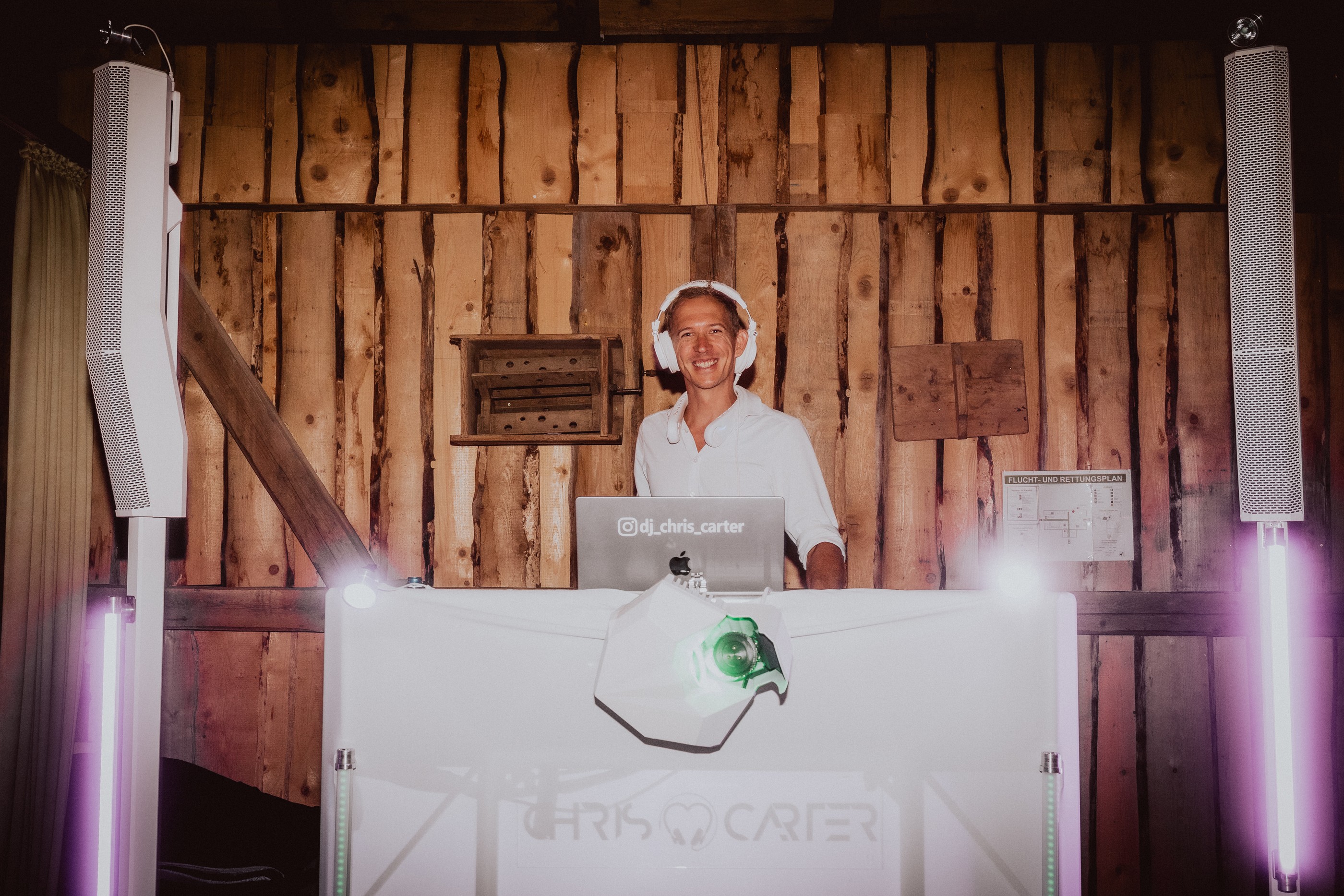 Chris Carter lacht mit weißen Kopfhörern hinter dem weißen DJ-Pult vor einer Holzwand, links und rechts weiße Säulenlautsprecher und leuchtende LightTubes